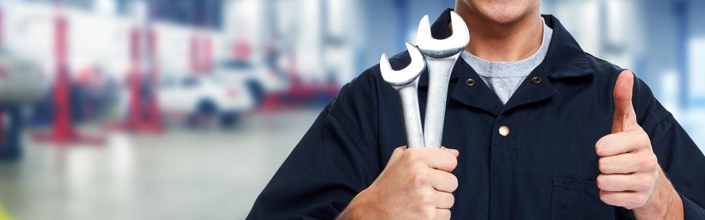 A mechanic in dark overalls holding wrenches and giving a thumbs-up in a repair shop.