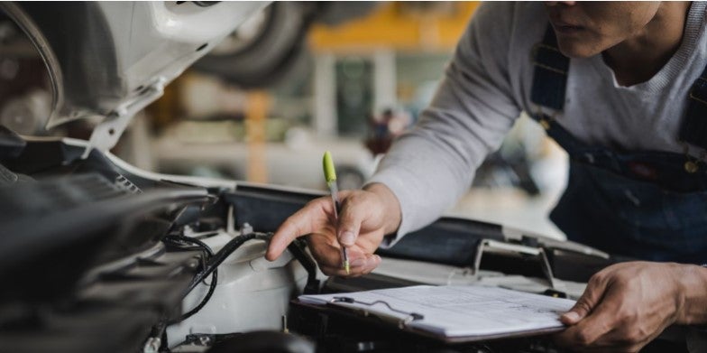 A mechanic inspecting an engine and making notes on a clipboard, while working on a vehicle.