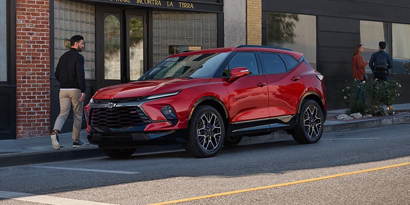 Front-three-quarter view of a bright red Chevrolet Blazer RS parked on a city street in front of brick storefronts, with pedestrians walking along the sidewalk nearby.