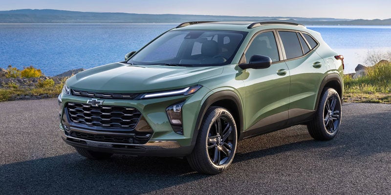 Front-three-quarter view of a sage green Chevrolet Trax parked on an asphalt overlook with a large body of water and hills in the background.