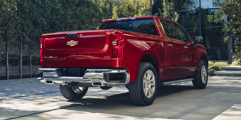 Rear-three-quarter view of a red Chevrolet Silverado pickup truck parked in a paved driveway next to a modern building with glass windows and surrounding greenery.