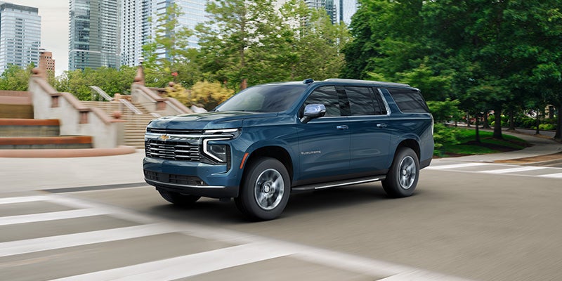 Front-three-quarter view of a dark blue Chevrolet Suburban driving on a city street past modern buildings.