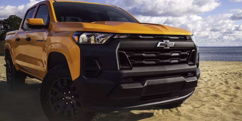 Front-three-quarter view of a bright yellow Chevrolet Colorado truck parked on a sandy beach near the water.