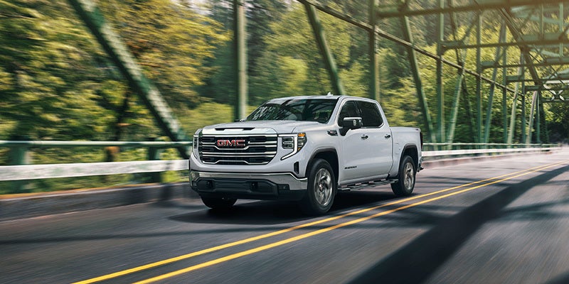 White GMC Sierra 1500 pickup truck driving across a steel bridge surrounded by lush greenery.