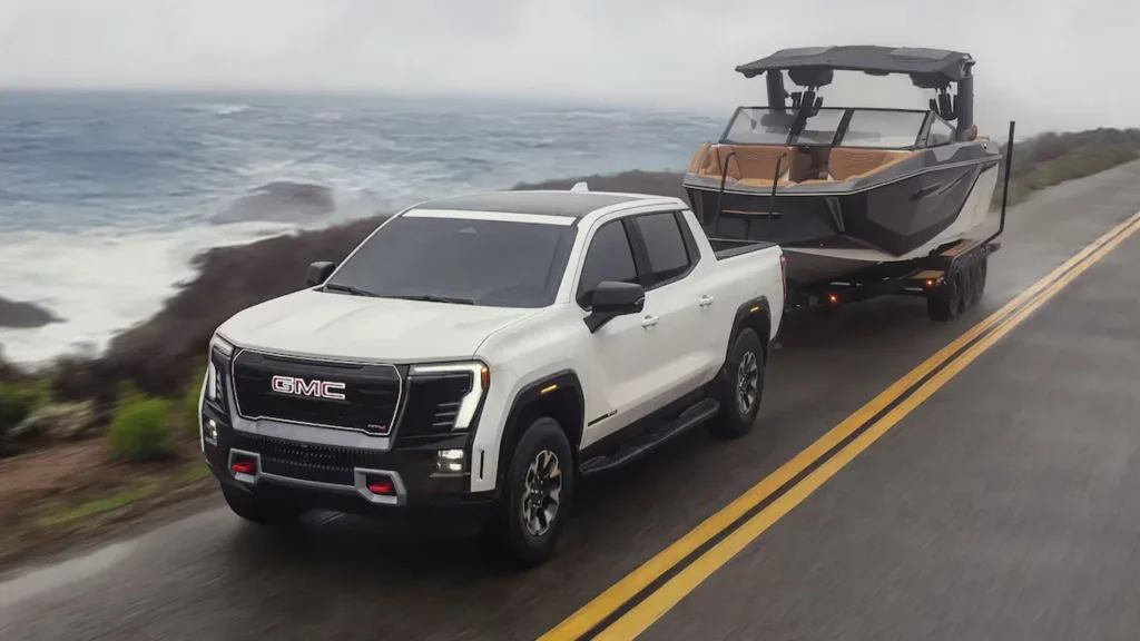 A white 2025 GMC Sierra EV Denali Edition 1 towing a luxury boat along a coastal highway, highlighting its electric power and premium towing strength.
