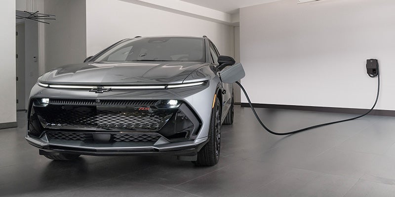 A sleek, silver electric Chevrolet Equinox EV is parked in a modern garage, plugged into a wall-mounted charger. The setting conveys innovation and eco-friendliness.