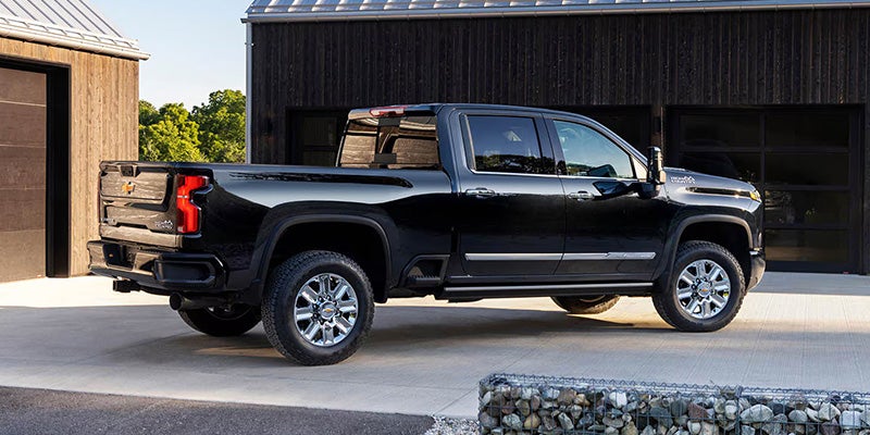A black pickup Chevrolet Silverado HD truck is parked on a light-colored driveway near a modern wooden building. The truck's polished surface reflects sunlight, conveying a sleek, robust appearance.