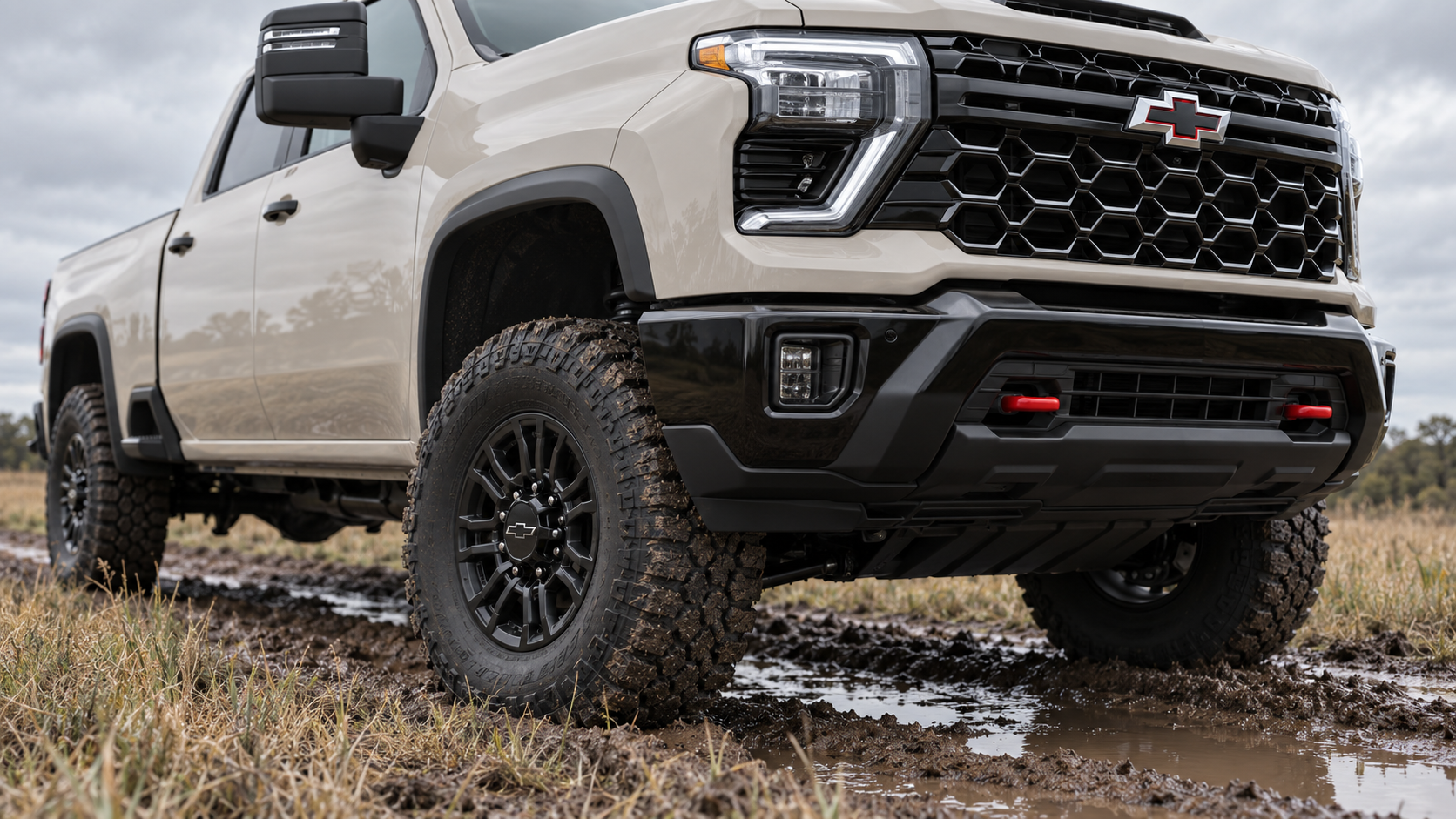 Low front corner of a 2026 Chevrolet Silverado 2500 HD ZR2 in White Sands showing the 35-inch mud-terrain tire, black off-road wheel, red recovery hooks and skid plate