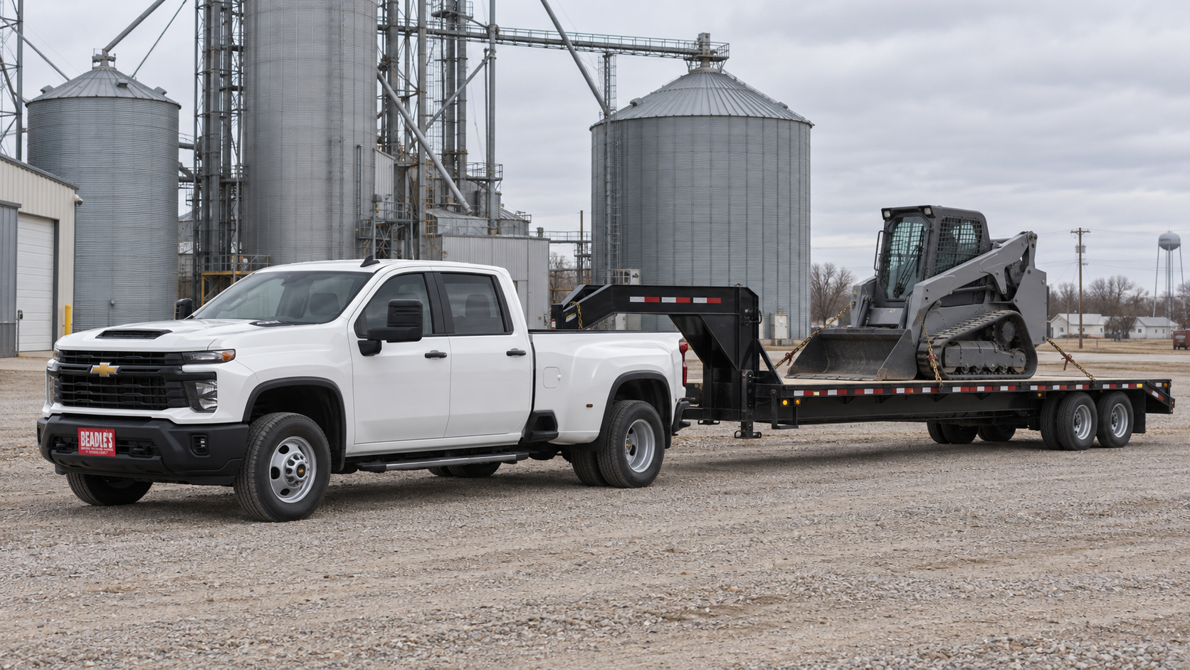 Chevrolet Silverado 3500 HD Work Truck dually hauling a compact loader on a gooseneck deck at a grain elevator in a small South Dakota town