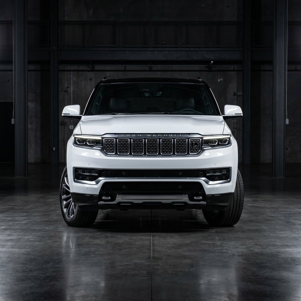 Front-facing view of a white Grand Wagoneer luxury SUV parked in a dark industrial studio. (Representative image for illustration purposes only. Actual vehicle may vary based on trim and configuration.)