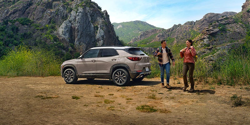 A beige 2026 Chevrolet Trax parked in a mountainous landscape, with two people walking nearby, enjoying an outdoor adventure.