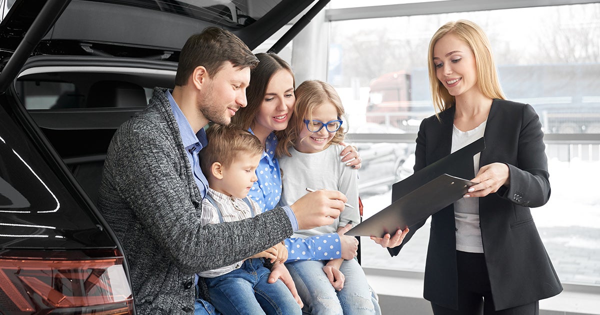 A family sitting in the back of a new vehicle in the car dealership signing paperwork with the car salesperson.