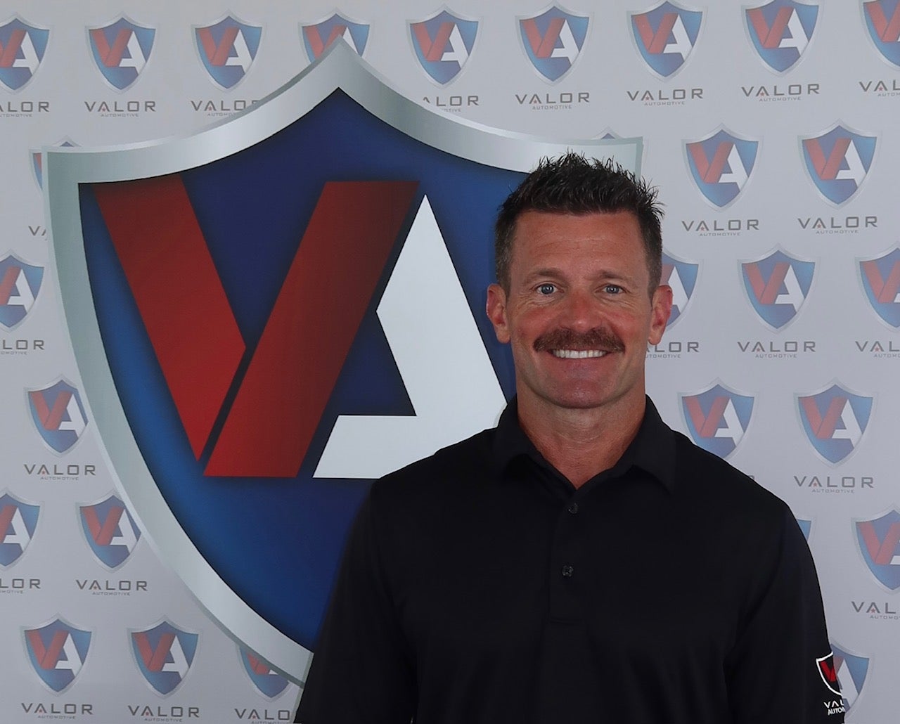 Smiling man with a mustache wearing a black polo shirt in front of a Valor Automotive logo backdrop.