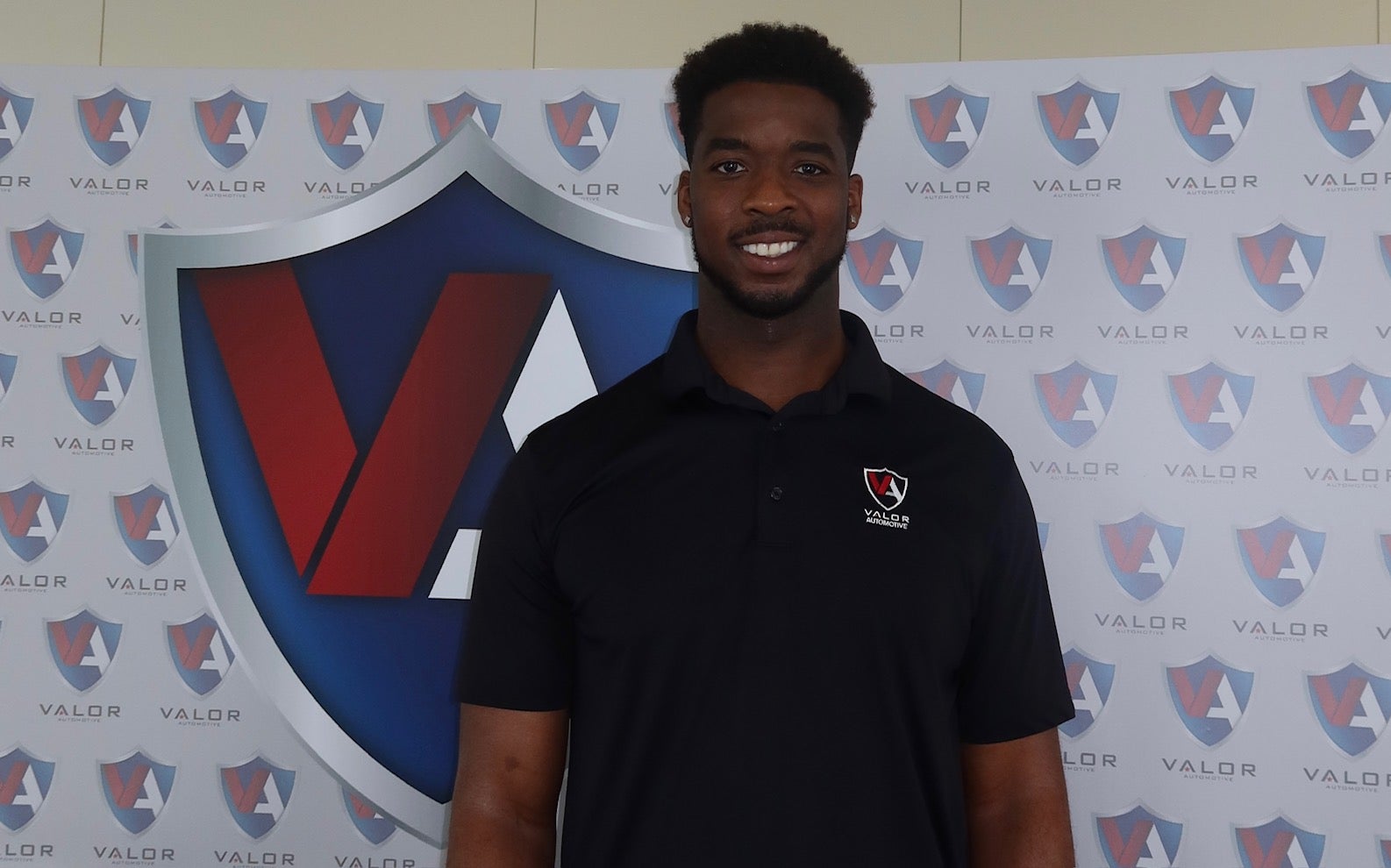 A smiling man in a black polo shirt standing in front of a Valor Automotive branded backdrop.