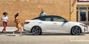 Side view of a white 2026 Nissan Sentra parked on a city street with three pedestrians walking nearby against a brick building background.