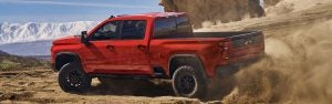Red 2026 Chevrolet Silverado 2500 driving on sandy terrain kicking up dust.
