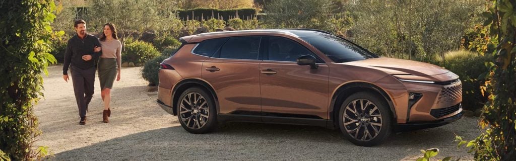 Couple walking beside a brown SUV on a scenic garden driveway.
