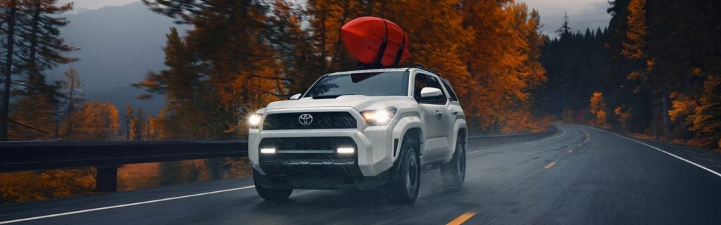 White Toyota 4Runner driving on a wet road with a red kayak on top, surrounded by autumn trees.