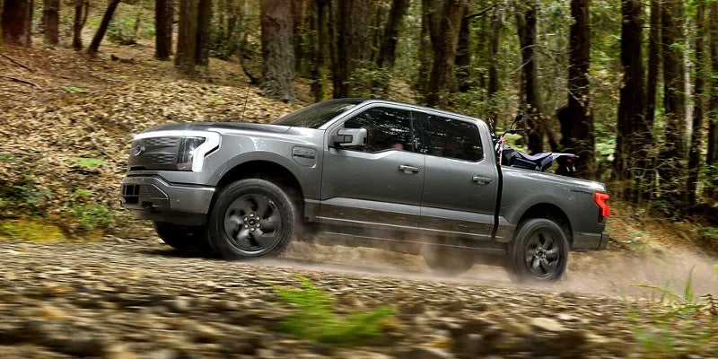 An all-new silver 2025 Ford Lightning hauling a motorcycle in the bed up a back road trail on a mountain. - Franklin Ford Franklin, MA