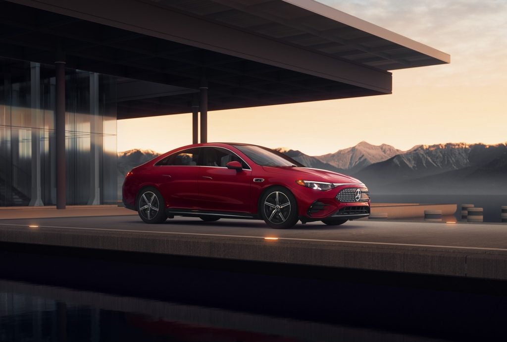 Red all-electric 2026 Mercedes-Benz CLA parked near a modern building with mountains in the background, highlighting luxury sedan styling.