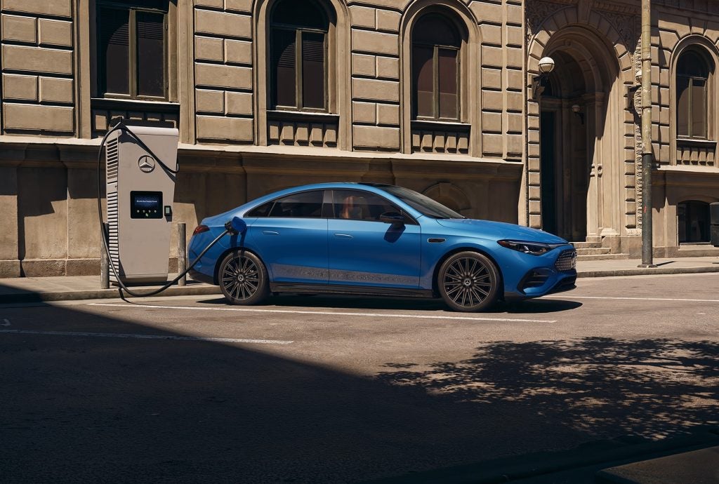 Blue all-electric 2026 Mercedes-Benz CLA charging at an EV station beside a city building.