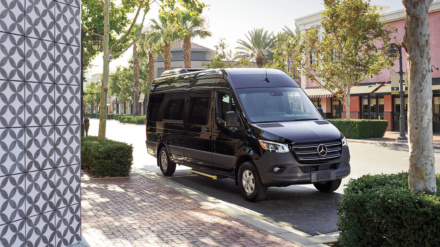 Black Mercedes-Benz Sprinter Van parked on a quiet city street with shops and trees in the background.