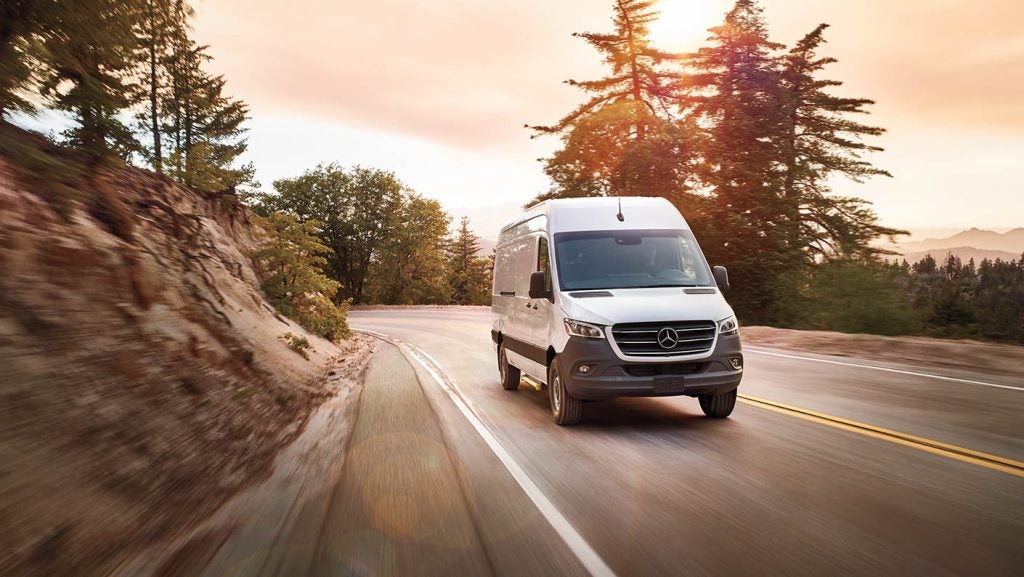 White Mercedes-Benz Sprinter Van driving on a curved mountain road at sunset with trees and cliffs surrounding the route.