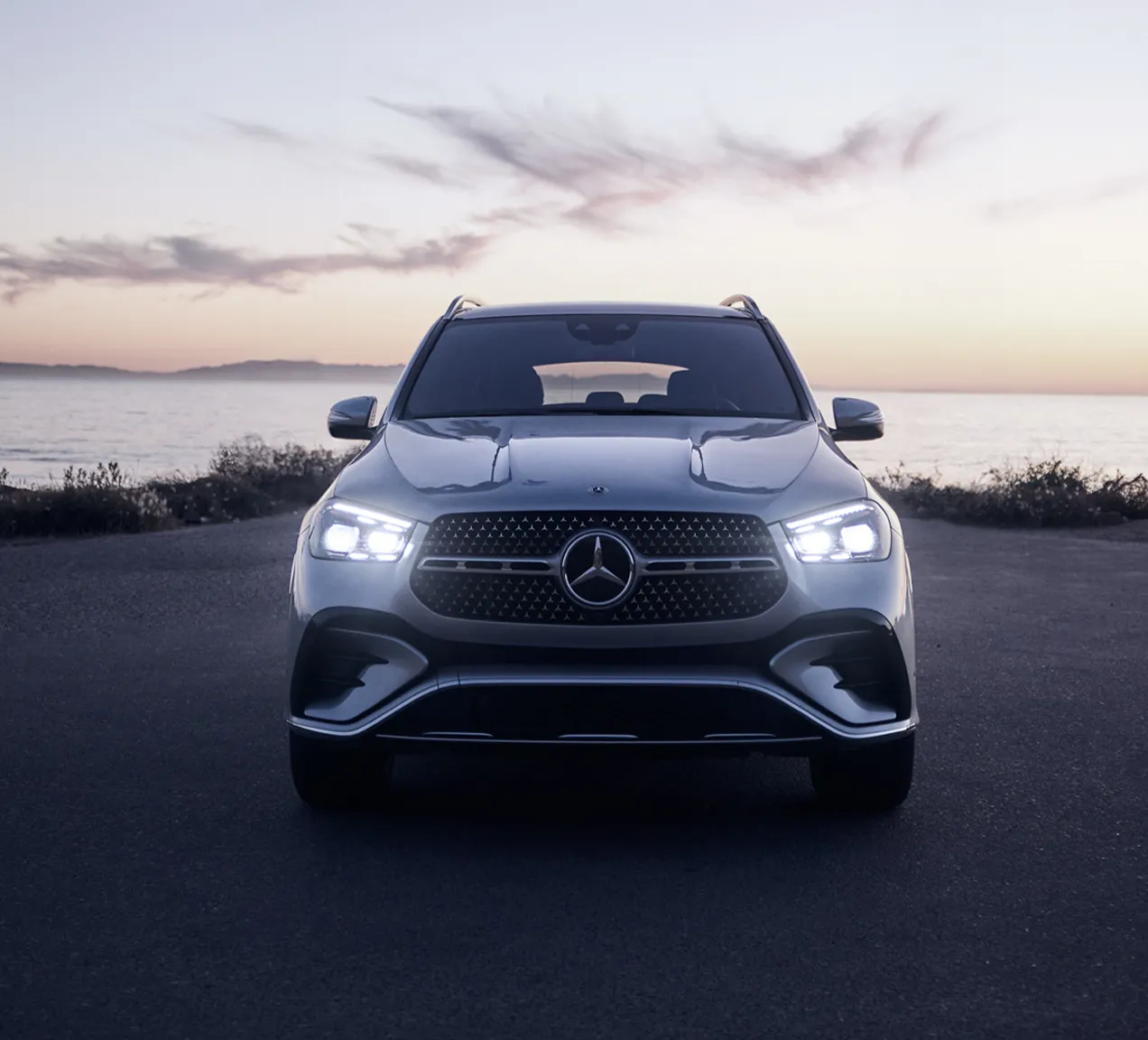 Front view of a 2026 Mercedes-Benz GLE parked by the ocean at sunset, showcasing its illuminated LED headlights and sleek grille, emphasizing its capability for enjoyable winter driving.