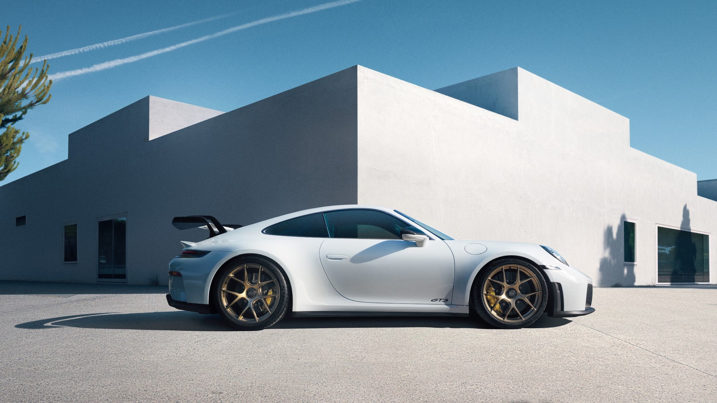 Side profile of a white Porsche 911 parked in front of a modern white building, representing Porsche’s next 911 and pure driving pleasure.