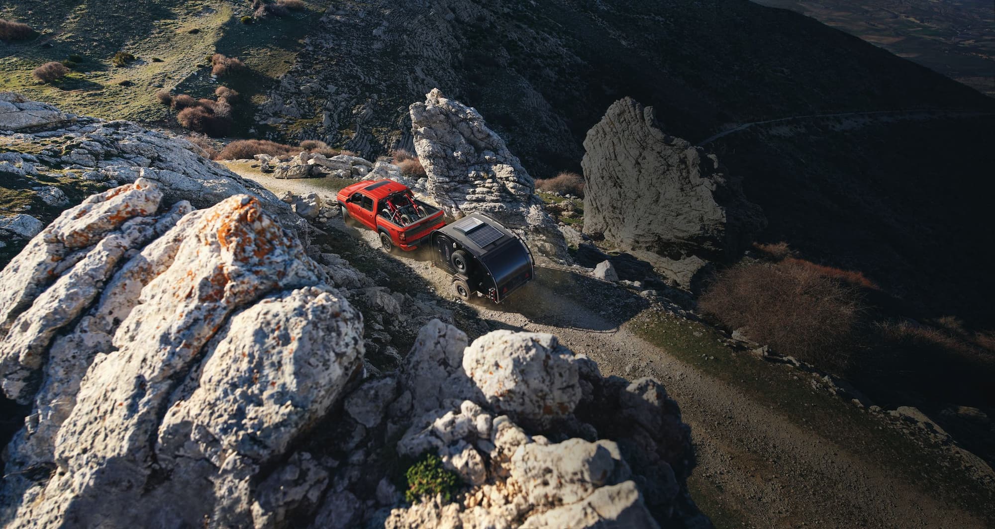 Red Nissan Frontier towing a black camper trailer on a rocky mountain off-road trail