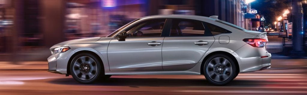 A side-profile view of a silver Honda Civic sedan driving past blurred city storefronts and streetlights at night.