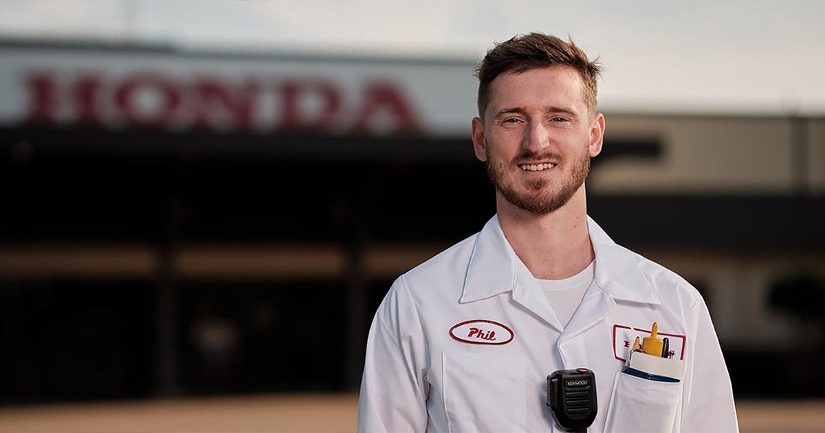 White-uniformed Honda technician smiling in front of dealership, wearing name tag and radio, Priority Honda Roanoke service team