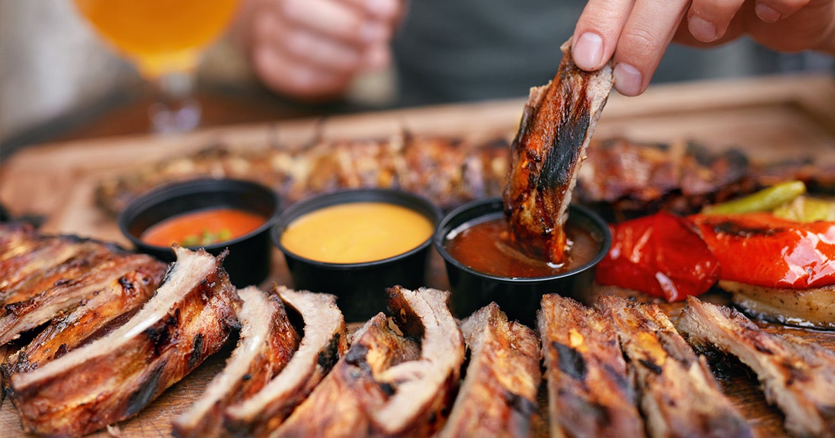 Grilled ribs with barbecue sauces, a hand dipping a rib into sauce, roasted red peppers on a wooden board
