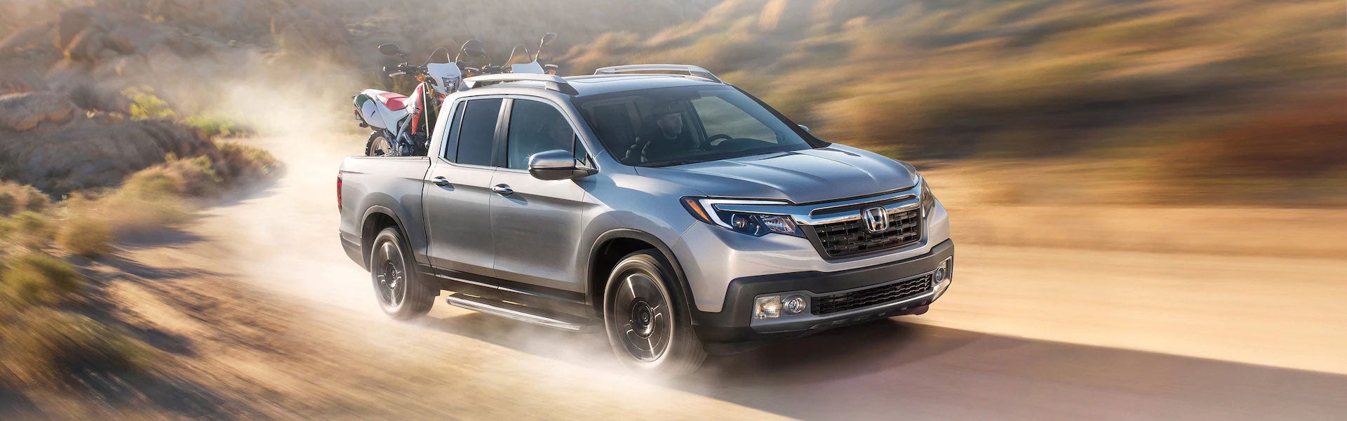 Silver Honda Ridgeline pickup truck driving on dusty off-road trail, carrying motorcycle in truck bed | Priority Honda Roanoke SUV crossover
