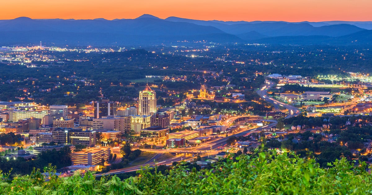 Aerial view of Roanoke, Virginia at twilight, showing downtown skyline with mountains in background and Priority Honda dealership lights visible