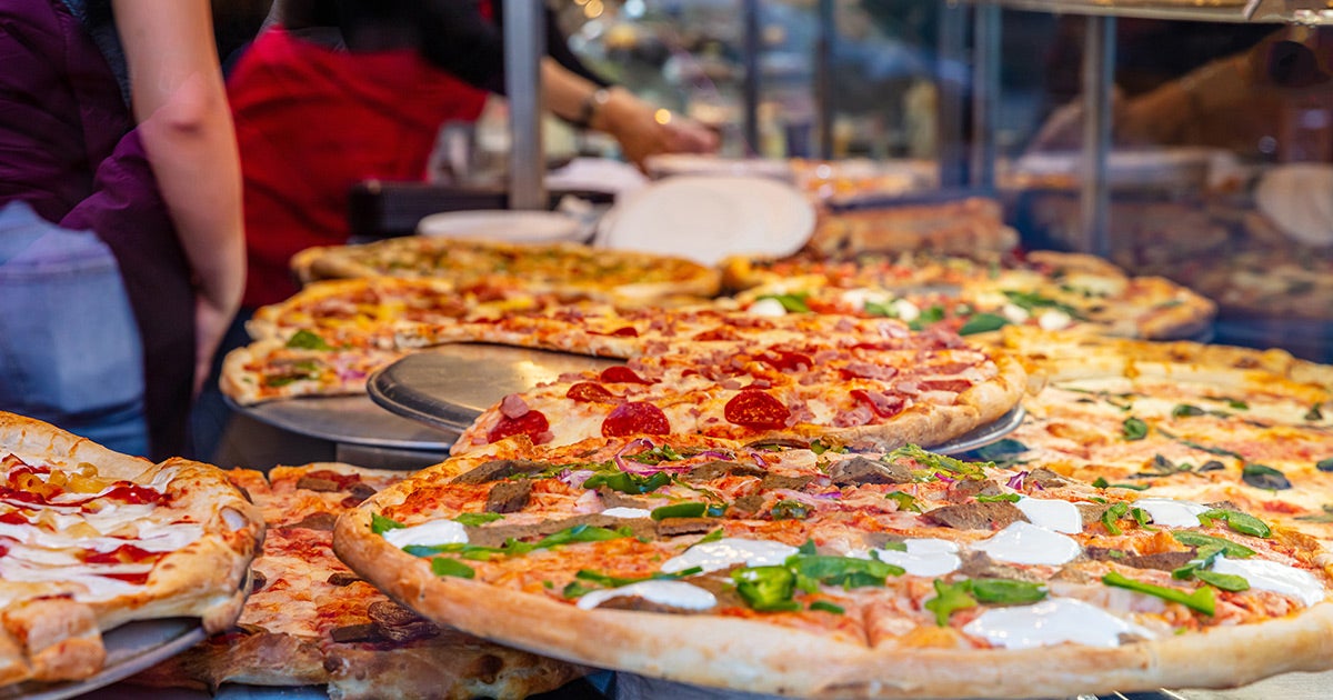 Multiple freshly made pizzas with various toppings like pepperoni, jalapeños, and mozzarella cheese, displayed at a pizza counter or restaurant with staff visible in the background.