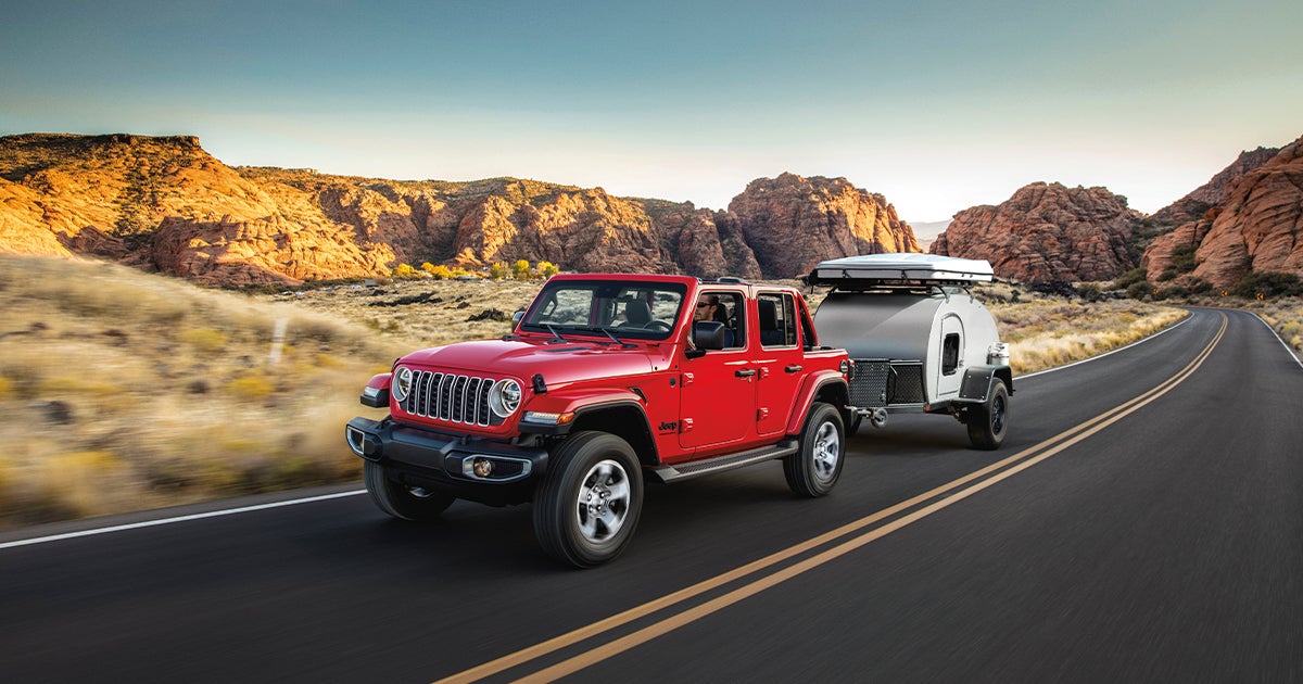 A red Jeep Wrangler towing a trailer on the road in a desert.
