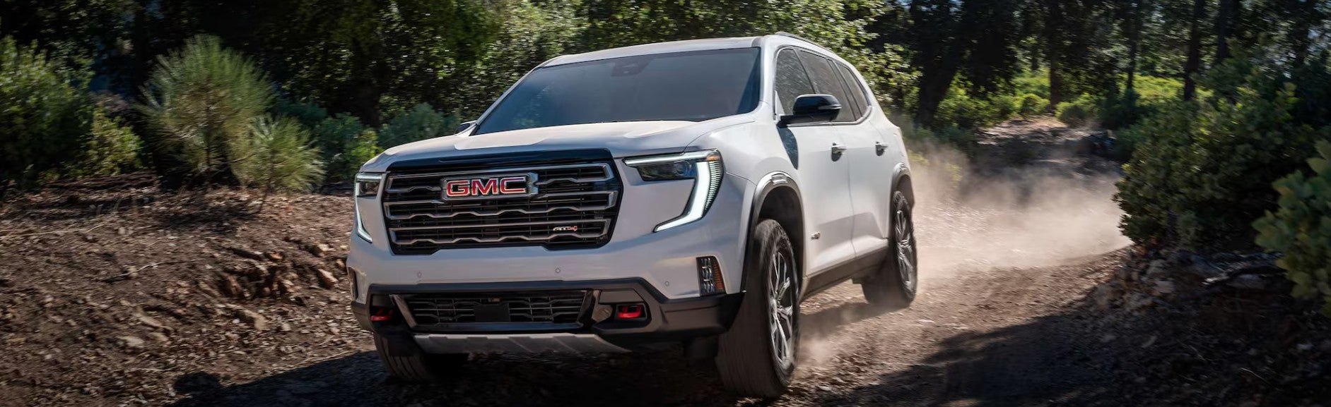 White 2027 GMC Acadia driving on a dirt road in the woods