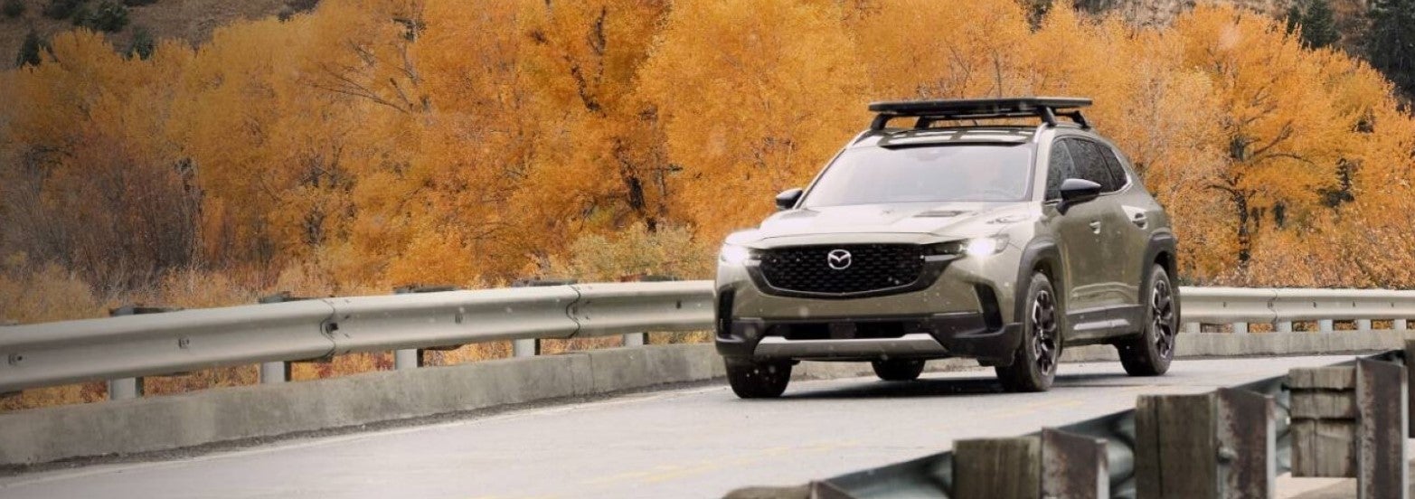 A new Mazda CX-50 drives on a bridge with orange fall trees in the background, making this Mazda a perfect travel companion. Shop for your new Mazda SUV at Mazda of Spartanburg.