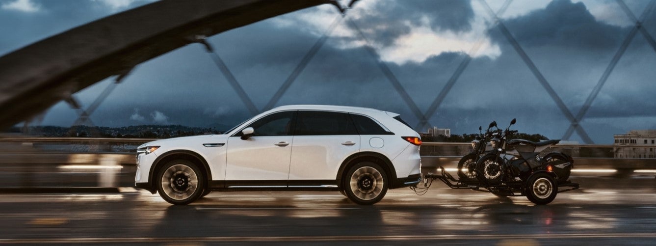 The new Mazda CX-90 is shown driving over a bridge whilst towing two motorcycles, making this SUV a perfect travel SUV. Shop for you new Mazda CX-90 at Mazda of Spartanburg.