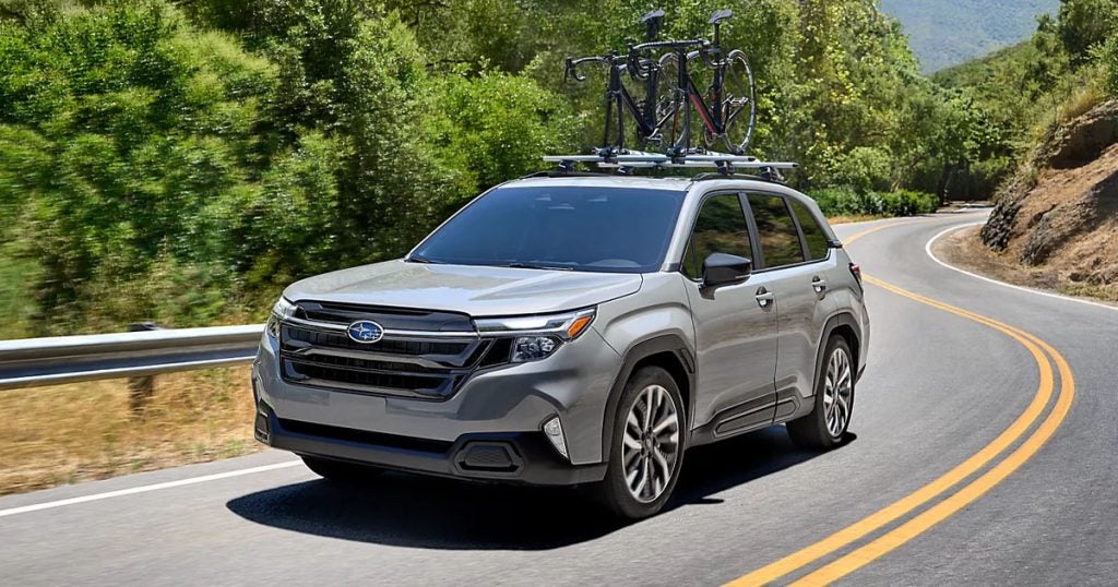 2025 Subaru Forester Touring trim in River Rock Pearl with the bike rack accessory and bikes on top driving on the highway up a mountain.