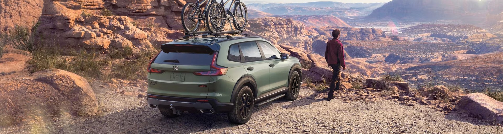A green Honda CR-V Hybrid with two bikes on the roof bike attachment parked by the edge of a cliff while a man looks over the horizon.
