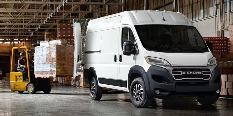 A white RAM Promaster in a warehouse. The back doors are open as a forklift driver loads a pallet into the back.