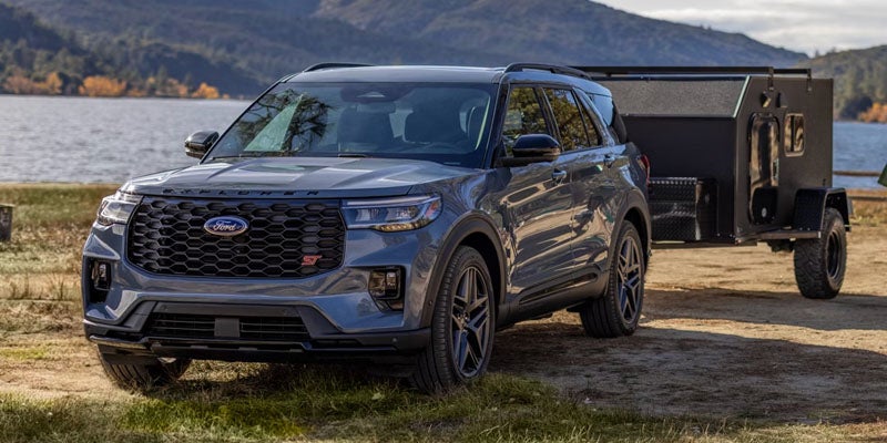 Blue Ford SUV towing a black enclosed trailer parked near a lake with mountains in the background, showcasing its towing capability and rugged outdoor design.