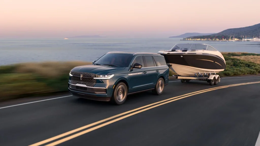 The 2025 Lincoln Navigator® Reserve in a rich green finish, towing a sleek black-and-white boat along a coastal highway at sunset.