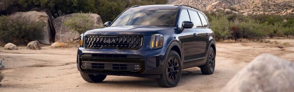 A Kia Telluride SUV parked on a rugged dirt path with rocks and shrubs in the background.