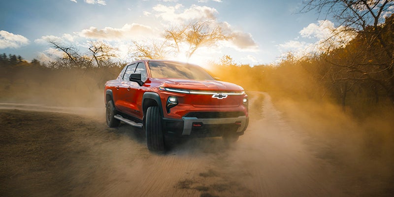 Orange Chevrolet Silverado EV driving through a dusty off-road trail with motion and trees in the background.