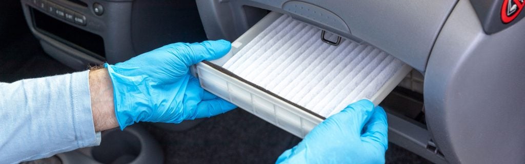 This image shows a person wearing blue gloves, removing a clean cabin air filter from a compartment in a car's dashboard, likely for maintenance or replacement.
