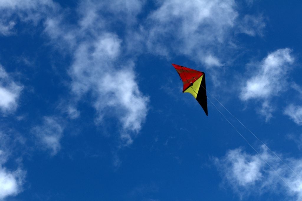 A black, red, and yellow kite flying high against a blue and white cloud sky.