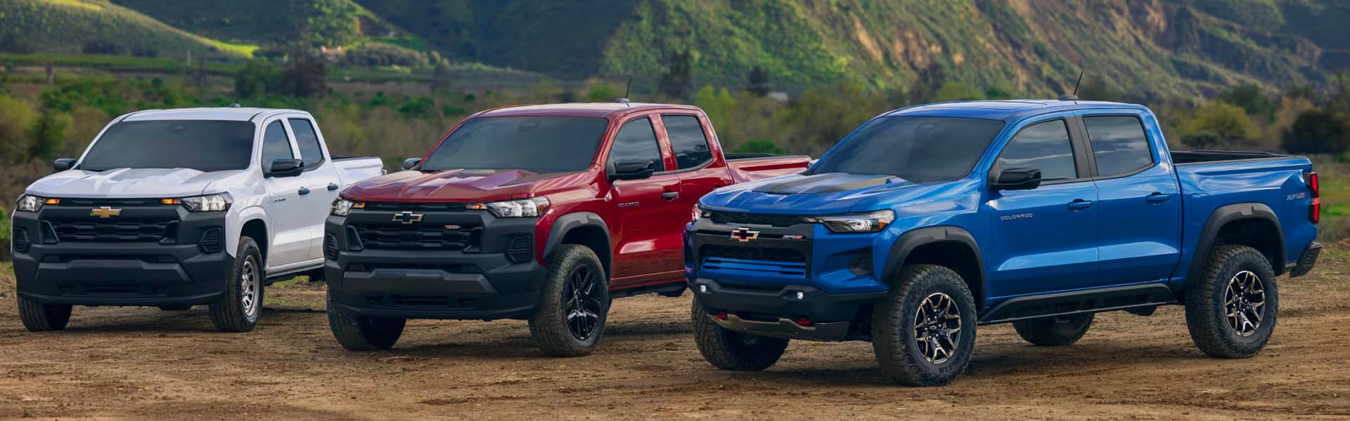 Front view of three white, red, and blue Chevy Colorado trucks lined up with rolling hills in the background